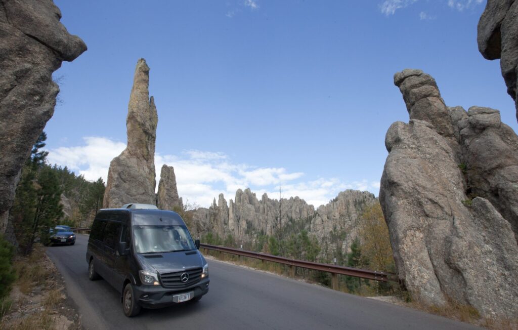 Road with towering rock spires on both sides and a dark van in the foreground, blue sky above, likely a scenic park area.